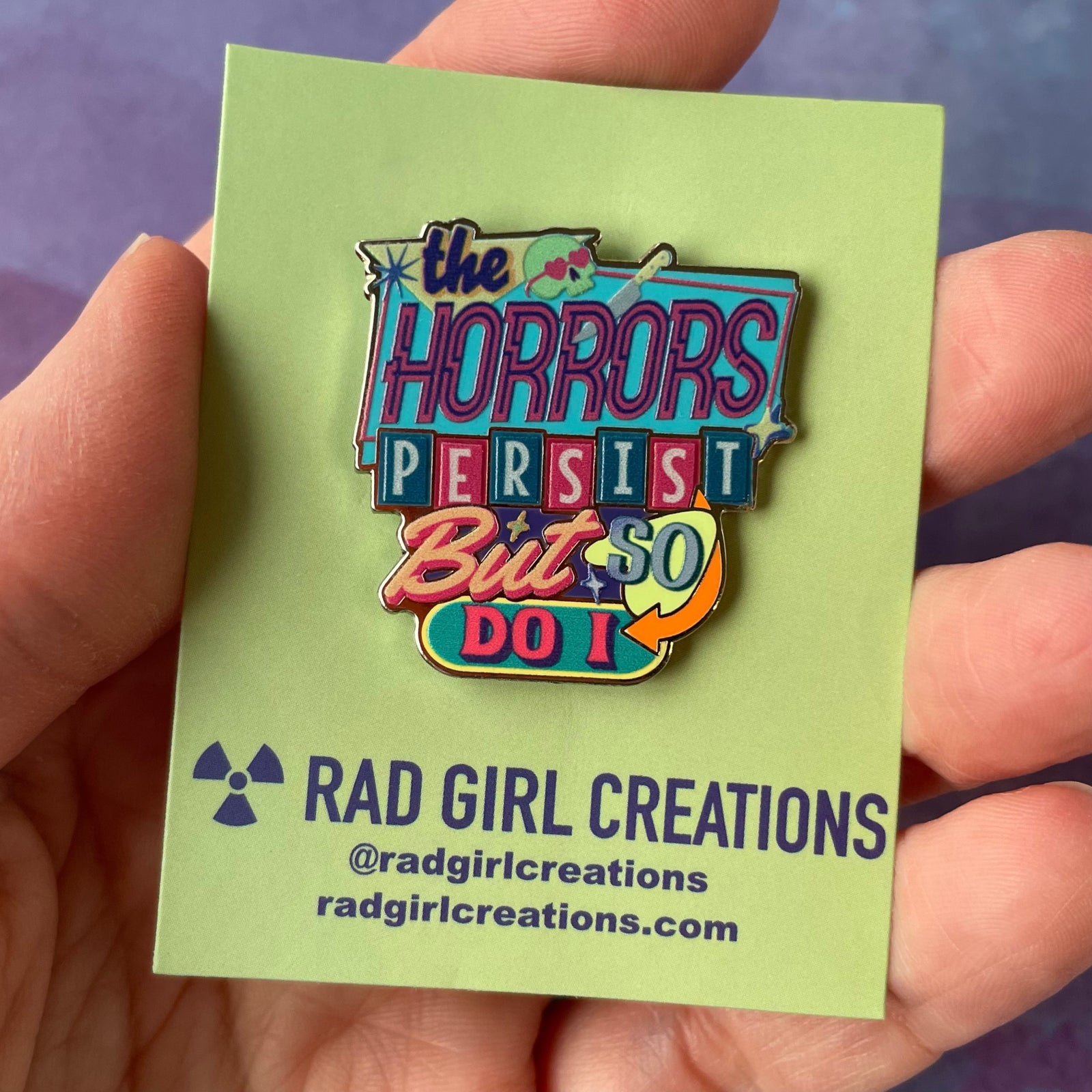 Colorful enamel pin with text 'the HORRORS PERSIST But SO DO I' on a gray background