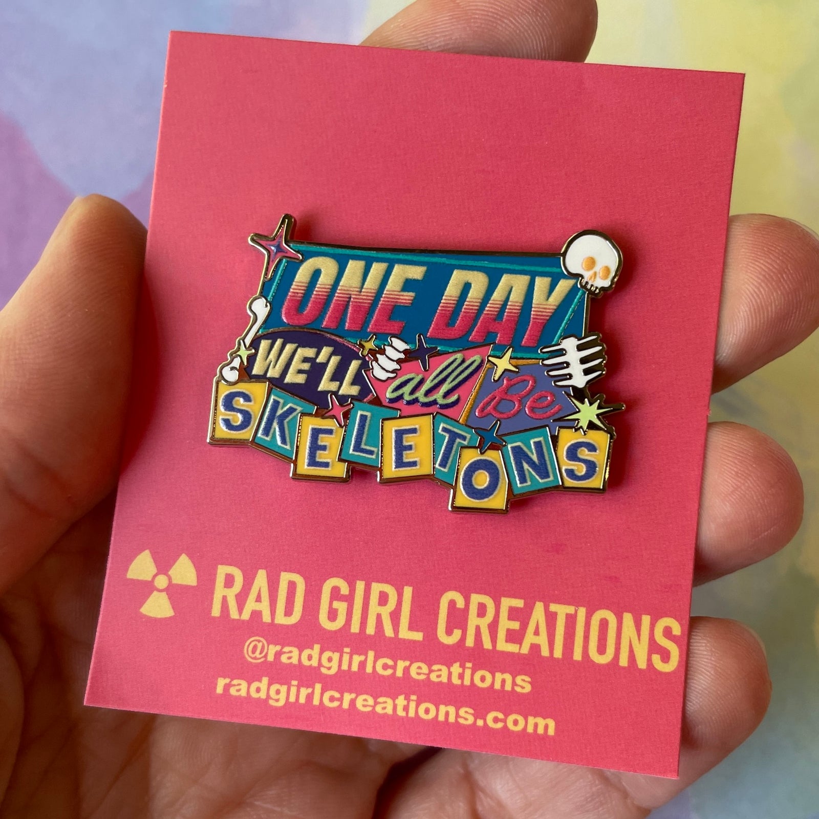 Colorful enamel pin with text 'One day we'll all be skeletons' on a gray background