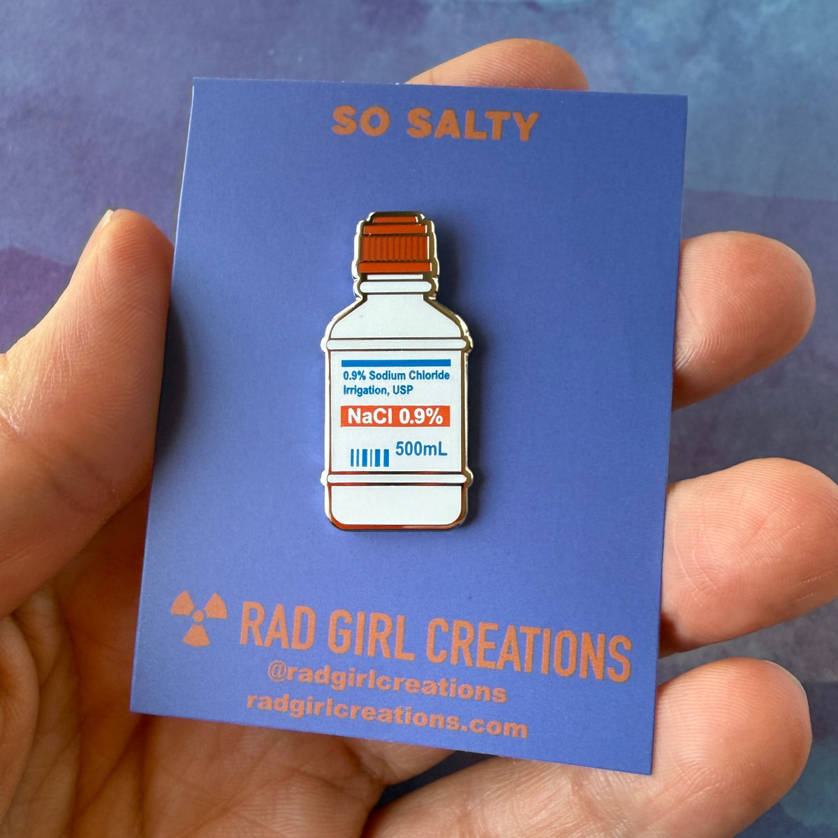 Photograph of an enamel pin shaped like a 500mL 0.9% Sodium Chloride irrigation bottle with a medical label.