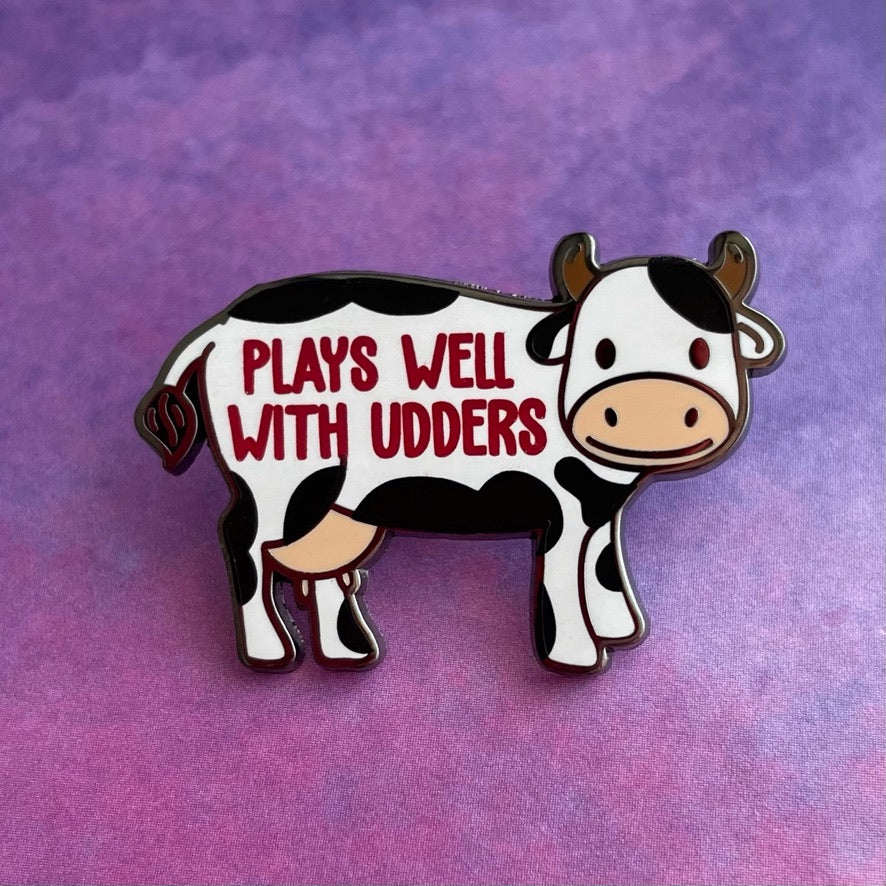 Cow-shaped pin with 'PLAYS WELL WITH UDDERS' text on a gray background