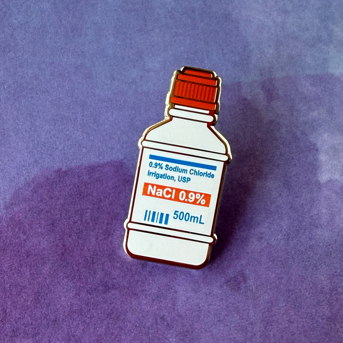 Photograph of an enamel pin shaped like a 500mL 0.9% Sodium Chloride irrigation bottle with a medical label.