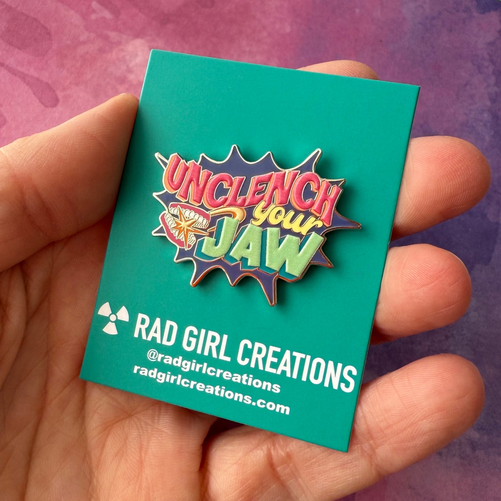 Colorful enamel pin with 'Unclench your Jaw' text on a gray background