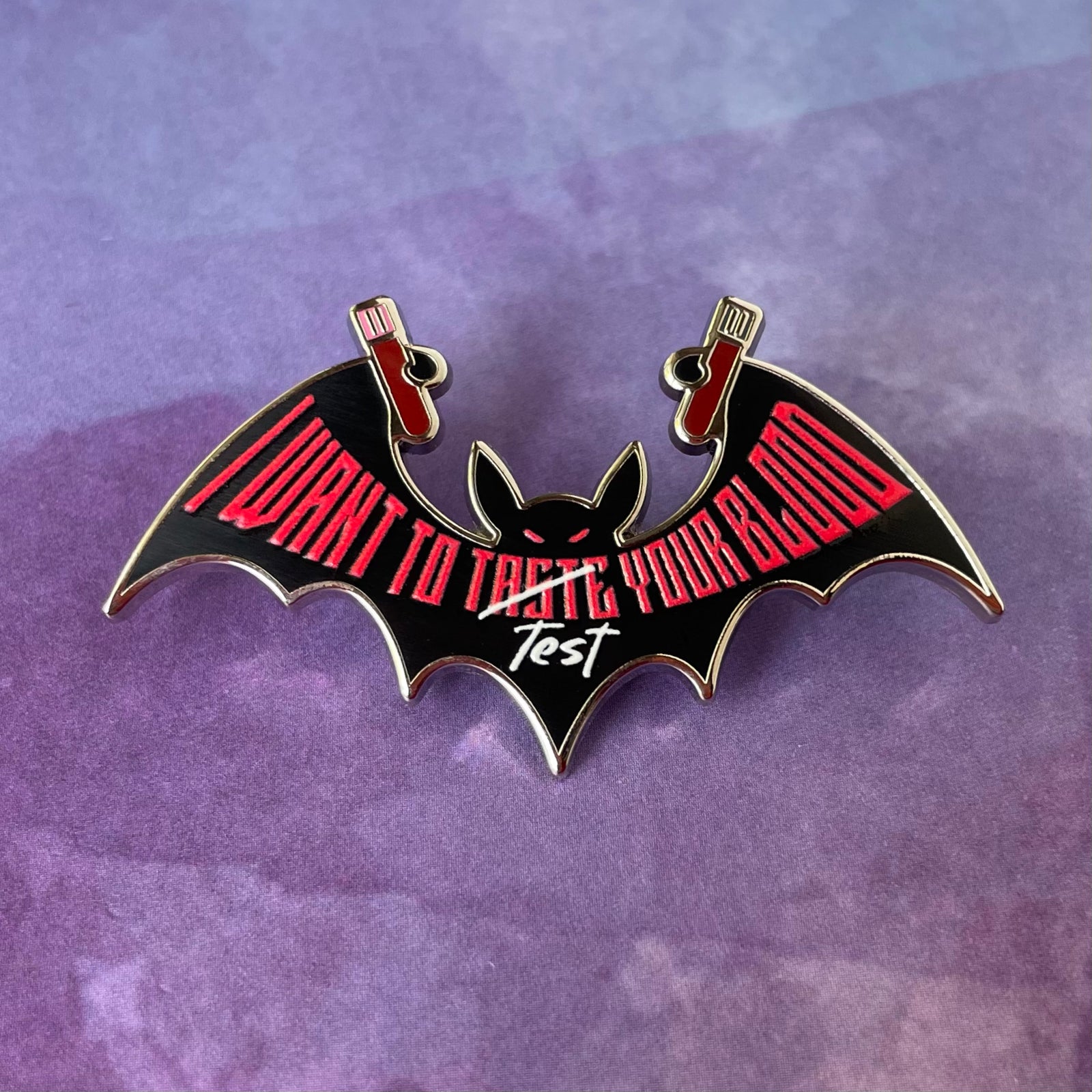 Bat-shaped enamel pin with 'I want to test your blood' text on a gray background