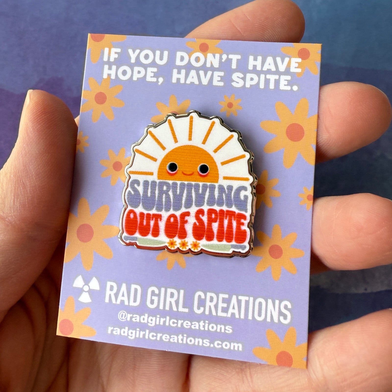 Enamel pin with a sun design and text 'Surviving Out of Spite' on a gray background