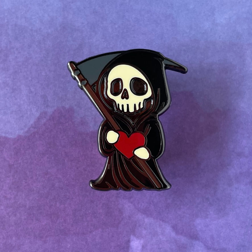 Pin with cartoon-style grim reaper holding a red heart on a light gray background